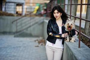 ragazza gitana bruna con cane yorkshire terrier posato contro ringhiere in acciaio. indossa il modello su giacca di pelle e t-shirt con ornamento, pantaloni. foto