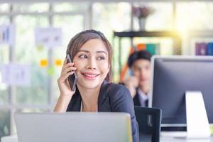 allegra imprenditrice utilizzando il telefono in ufficio. ragazza carina eccitata che utilizza smartphone e collega sfocato seduto sullo sfondo foto