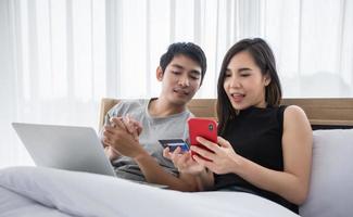 coppia asiatica che acquista online e paga con carta di credito su laptop e telefono cellulare a casa, coppia felice a casa che naviga in rete a letto foto