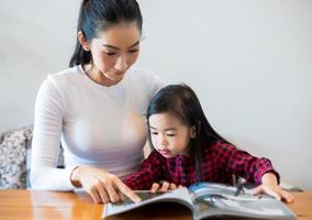 una madre asiatica sta insegnando a sua figlia a leggere un libro durante la pausa del semestre sul tavolo del soggiorno e a bere il latte freddo sul tavolo di casa. concetti e attività educative della famiglia foto