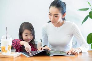 una madre asiatica sta insegnando a sua figlia a leggere un libro durante la pausa del semestre sul tavolo del soggiorno e a bere il latte freddo sul tavolo di casa. concetti e attività educative della famiglia foto