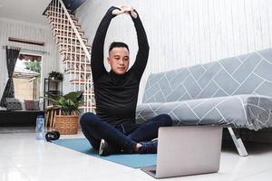 l'uomo asiatico fa esercizi di stretching al chiuso a casa usando il laptop come guida foto