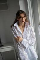 attraente bruna sexy in camicia bianca in posa provocatoria, vicino a una finestra, girato in studio. ritratto di donna sensuale con i capelli lunghi, nella classica scena del boudoir, guardava fuori dalla finestra foto