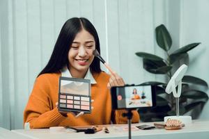 giovane donna asiatica che vlogging sui prodotti cosmetici per la cura della pelle sul tavolo con la sua videocamera e mostra l'uso del prodotto e le recensioni per il suo canale blog online. foto