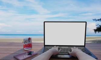 un computer o laptop posizionato su un tavolo di legno, uno smartphone nelle mani di una persona. lo schermo è bianco vuoto. sfondo di mare e spiaggia. allenarsi, vacanza al mare, rendering 3d foto