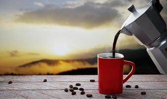 versare il caffè o l'espresso appena preparato dalla moka in una tazza da caffè rossa. caffè caldo in una tazza posizionata sul ripiano del tavolo o sul balcone di legno. vista sulle montagne mattutine, sole mattutino. rendering 3D foto
