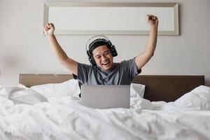 uomo asiatico felice in cuffia che esamina il computer portatile con gesto vincente che esprime eccitato sul letto foto