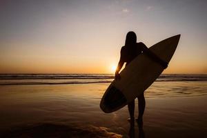 sagoma di bella donna sexy surfista con tavola da surf sulla spiaggia di sabbia al tramonto. sport acquatici. il surf è uno stile di vita sano e attivo. vacanze estive. foto