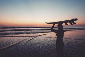 sagoma di bella donna sexy surfista con tavola da surf sulla spiaggia di sabbia al tramonto. sport acquatici. il surf è uno stile di vita sano e attivo. vacanze estive. effetto colore retrò. foto