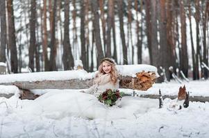 la graziosa sposa di aspetto slavo con una corona tiene un bouquet, si siede accanto al tronco nella foresta innevata. cerimonia di nozze d'inverno. foto