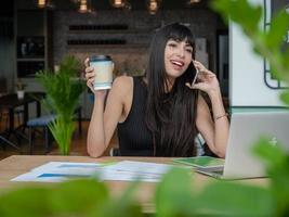 attraente donna d'affari caucasica che utilizza un telefono cellulare ed effettua una chiamata mentre è seduto sul tavolo con un documento informativo e lavora in un ufficio moderno foto