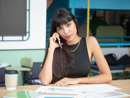 attraente donna d'affari caucasica che utilizza un telefono cellulare ed effettua una chiamata mentre è seduto sul tavolo con un documento informativo e lavora in un ufficio moderno foto