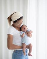 felice madre asiatica che tiene un adorabile neonato nella camera dei bambini, donna madre che abbraccia il suo neonato in piedi vicino alla finestra foto