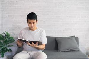 ritratto di gioioso uomo asiatico che legge un libro mentre è seduto sul pavimento nel suo soggiorno. uomo asiatico relex libro di lettura sorriso al divano in casa. foto