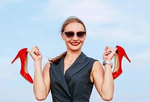 ritratto di giovane donna bianca alla moda in tuta, occhiali da sole, in piedi con le scarpe lucide con i tacchi alti nelle sue mani all'aperto foto