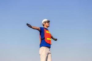 giovane di successo con le braccia spalancate, felice caposquadra ingegnere in elmetto protettivo e giubbotto di sicurezza parla su sfondo blu cielo foto