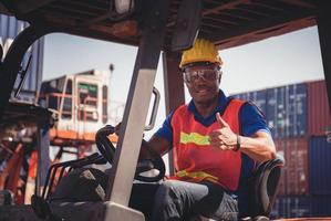 uomo lavoratore in elmetto protettivo e giubbotto di sicurezza seduto in impilatori di contenitori sorridendo con i pollici in su in segno di successo foto