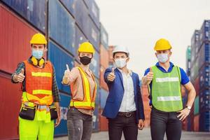 squadra di operai e ingegneri che indossano una maschera protettiva contro il coronavirus con i pollici in su ai container foto