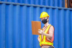 ingegnere che indossa una maschera protettiva contro il coronavirus, caposquadra donna con elmetto protettivo e giubbotto di sicurezza che tiene la lista di controllo degli appunti al carico dei container foto