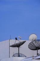 3 antenne paraboliche con antenna tv in cima al vecchio edificio grigio su sfondo blu cielo limpido in cornice verticale foto