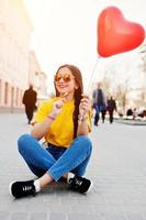 giovane ragazza seduta per le strade della città con lecca-lecca e palloncino cuore a portata di mano, indossare t-shirt gialla, jeans e occhiali da sole. foto
