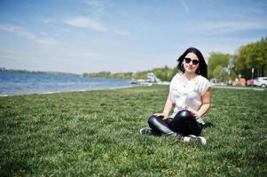 ragazza bruna su pantaloni di pelle da donna e camicetta bianca, occhiali da sole, seduta su erba verde contro la spiaggia del lago. foto