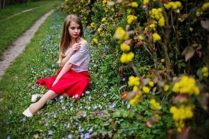 ritratto di sitiing bella ragazza con labbra rosse al giardino di fiori di primavera su erba con fiori, indossare su abito rosso e camicetta bianca. foto