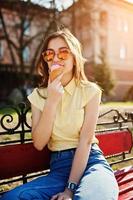 giovane ragazza seduta su una panchina con gelato a portata di mano, indossare t-shirt gialla, jeans e occhiali da sole. foto