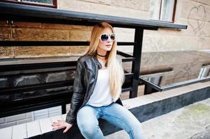 elegante donna bionda indossa jeans, occhiali da sole, girocollo e giacca di pelle in strada. ritratto di modello urbano di moda. foto