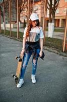 giovane ragazza urbana adolescente con skateboard, indossare occhiali, berretto e jeans strappati al campo sportivo del cortile. foto