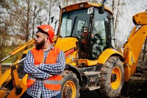 barba brutale lavoratore uomo vestito operaio edile in casco arancione di sicurezza contro traktor. foto