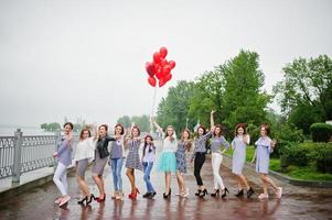 undici trecce damigelle dall'aspetto sorprendente con splendida sposa in posa con palloncini rossi a forma di cuore sul marciapiede contro il lago sullo sfondo. foto