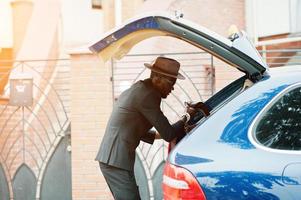 elegante uomo di colore con gli occhiali con cappello, indossare tuta con borsetta contro auto di lusso. ricco uomo d'affari afroamericano. foto