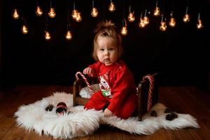 Una bambina di 7 mesi in un costume rosso di natale su uno sfondo di ghirlande retrò si siede su una pelliccia foto