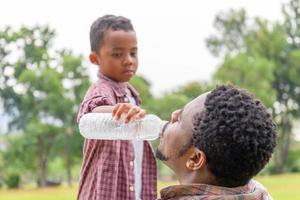 figlio che dà acqua a suo padre, papà afroamericano e figlio all'aperto, famiglia di felicità nei concetti del parco foto