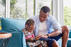 padre e figlia afroamericani allegri che giocano in soggiorno, bambina e papà carini seduti sul divano e che giocano sul tablet, concetti di famiglia di felicità foto