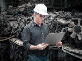 ingegnere meccanico caucasico di mezza età che utilizza la lista di controllo del computer portatile vecchio motore di auto usate nel magazzino del negozio di parti di macchinari. foto