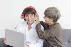 una coppia maschile con un uomo asiatico seduto su un divano con il loro amore reciproco e un altro che ascolta musica mentre suona il proprio laptop. foto