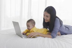 una mamma asiatica sta lavorando su un laptop a letto e con il suo bambino accanto al concetto di lavoro a casa. foto