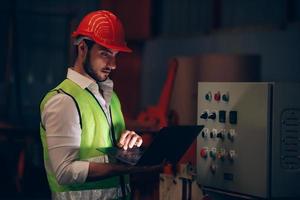 operaio di fabbrica che controlla l'impianto elettrico e il processo della macchina tramite laptop durante il lavoro straordinario in un cantiere industriale foto