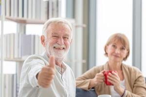 coppia senior dentro casa durante una pausa caffè, sorridente uomo anziano che mostra i pollici in su con in mano tazze di caffè foto