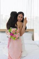 una sposa asiatica in abito da sposa bianco abbraccia la sua amica in abito rosa con un sorriso, poi mostra un anello di diamanti al dito sinistro e, dall'altra parte, tiene in mano un bellissimo mazzo di fiori. foto