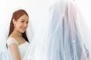 una sposa asiatica in un abito da sposa bianco sta sorridendo brillantemente davanti a un riflesso speculare. foto