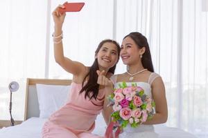 l'amica della sposa sta usando uno smartphone per fare un selfie di una sposa asiatica che tiene un mazzo di bei fiori in un abito da sposa nello spogliatoio sul letto e fa un piccolo segno con la mano del cuore. foto