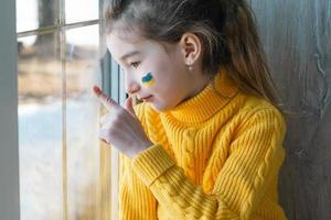 un bambino triste guarda la finestra con la bandiera dell'ucraina dipinta sulla guancia, preoccupazioni e paure. aiuti umanitari ai bambini, pace nel mondo, sicurezza. foto