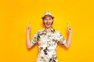 un giovane turista maschio felice isolato su uno sfondo giallo punta il dito indice verso un'area vuota. indossa un cappello, una camicia hawaiana. spazio per il testo. concetto - ricreazione, turismo, persone. foto
