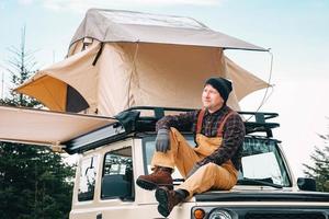 uomo avventuriero seduto sul tetto dell'auto con la tenda montata su di esso per lo stile di vita del campeggio di viaggio gratuito foto