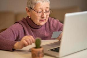 una donna anziana con i capelli grigi si siede davanti a un laptop con una carta di credito in mano, fa acquisti online. concetto di acquisto online foto