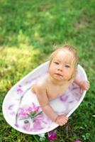 la ragazza felice del bambino prende un bagno di latte con i petali. bambina in un bagno di latte su sfondo verde. mazzi di peonie rosa. fare il bagnetto. igiene e cura dei bambini piccoli. foto