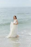 una giovane bella ragazza con un lungo vestito color latte cammina lungo la spiaggia e il molo sullo sfondo del mare. foto